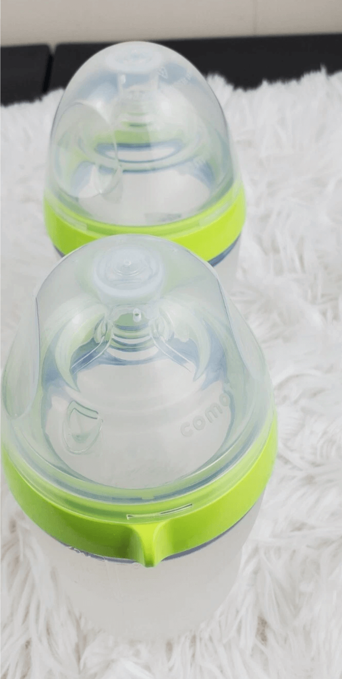 Two baby bottles with green accents on a textured white surface. Comotomo baby bottle double pack